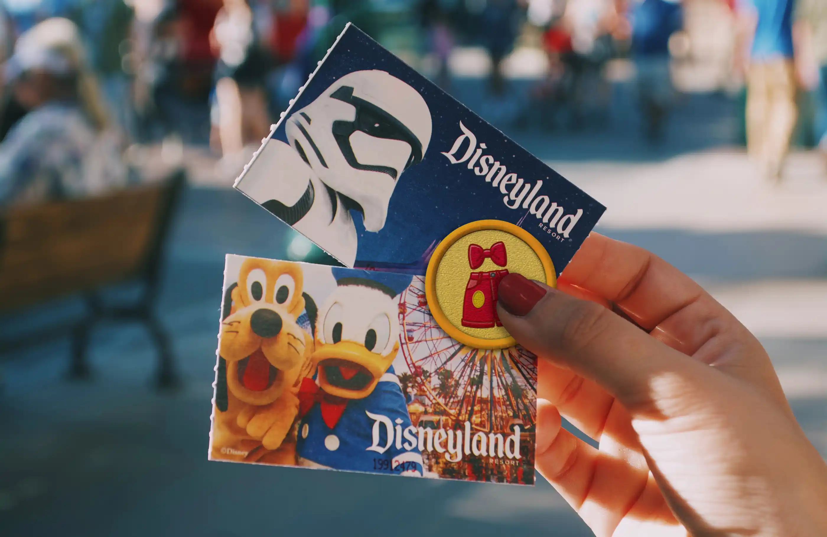 Hand holding small round patch and pair of Disneyland tickets.